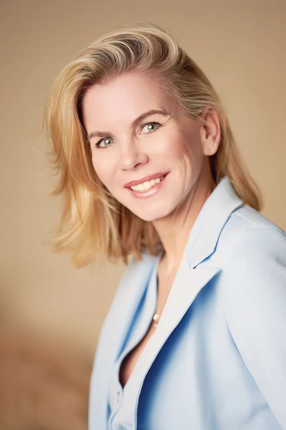 Lächelndes Businessportrait einer blonden Frau im hellblauen Blazer vor warmem Studiohintergrund.